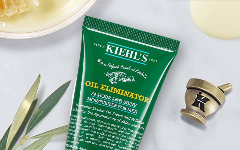 Bildbeschreibung: Eine grune Tube mit dem Text "Oil Eliminator" von Kiehl's liegt auf einer marmorartigen Oberflache, umgeben von kosmetischen Produkten und einer kleinen Schale mit einer unbekannten Substanz.