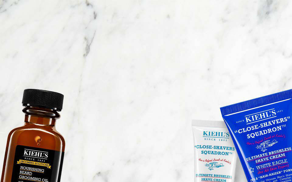 Alt-Text: Eine Anordnung von Kiehl’s Produkten auf einem marmorierten Hintergrund, einschließlich eines Gesichtsols, einer Rasiercreme und einer Aftershave-Creme.