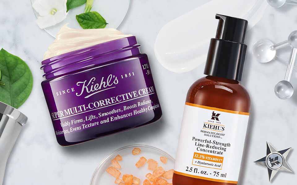 Eine helle, marmorierte Oberflache mit Hautpflegeprodukten, darunter eine lila Creme und ein braunliches Serum, umgeben von Pflanzenblattern und dekorativen Elementen.