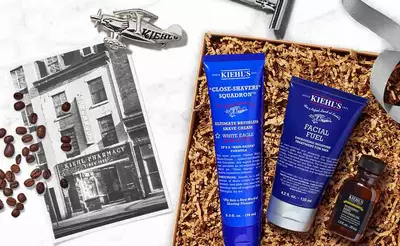 Geschenkset von Kiehl's mit Rasiercreme, Gesichtscreme und Vintage-Flugzeug, umgeben von Kaffeebohnen und Verpackungsmaterial.