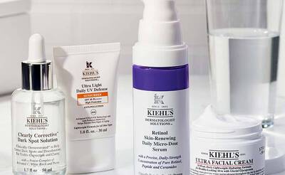 Vier Kiehl's Hautpflegeprodukte auf einer weißen Oberflache, darunter Seren und eine Tagescreme. Ein Glas Wasser im Hintergrund.