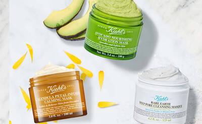 Drei Kiehl's Gesichtsmasken in verschiedenen Topfen, umgeben von Avocado und Blutenblattern.