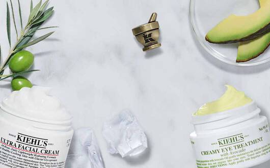 Zwei Tiegel von Kiehl's Gesichtspflegecreme auf einem marmorierten Tisch, umgeben von Avocado und Olivenzweigen.