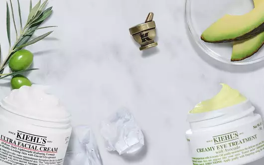 Zwei Tiegel von Kiehl's Gesichtspflegecreme auf einem marmorierten Tisch, umgeben von Avocado und Olivenzweigen.