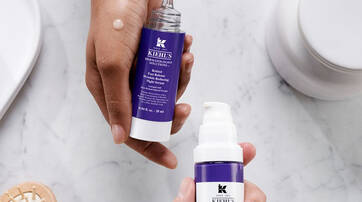 Deux flacons de serum de la marque Kiehl's, l'un avec un embout compte-gouttes et l'autre avec une pompe, poses sur une surface marbree. Une main tient l’un des flacons, tandis qu'une goutte de produit est visible sur l'autre.