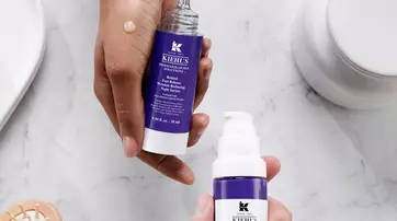 Deux flacons de serum de la marque Kiehl's, l'un avec un embout compte-gouttes et l'autre avec une pompe, poses sur une surface marbree. Une main tient l’un des flacons, tandis qu'une goutte de produit est visible sur l'autre.