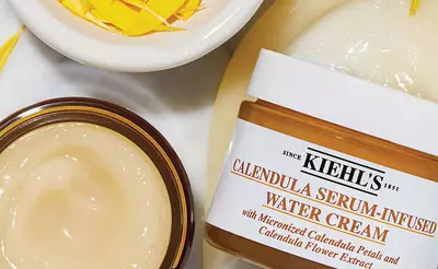Nahaufnahme von Kiehl's Calendula Wassercreme und einem Schalchen mit Ringelblumenblattern auf einem hellen Hintergrund.