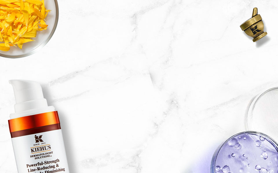 Alt-Text: Eine flache Anordnung von Kiehl's Hautpflegeprodukten auf einem marmorierten Hintergrund, einschließlich einer Tube mit Gesichtscreme, einer Flasche mit Lotion und frischen Krautern.