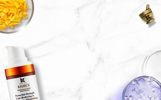Alt-Text: Eine flache Anordnung von Kiehl's Hautpflegeprodukten auf einem marmorierten Hintergrund, einschließlich einer Tube mit Gesichtscreme, einer Flasche mit Lotion und frischen Krautern.