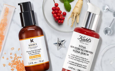 Zwei Hautpflegeprodukte von Kiehl's: ein Anti-Aging-Konzentrat und ein Hautstraffungsserum, umgeben von Krautern und Zutaten.