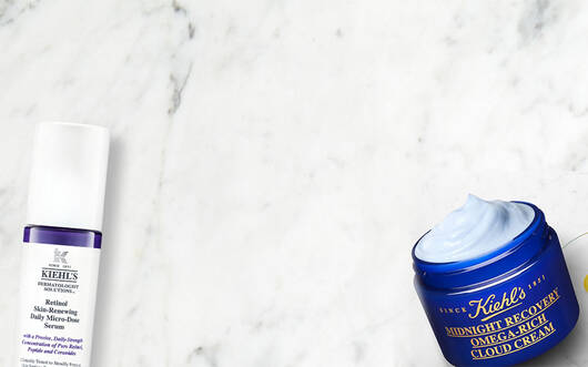 Alt-Text: Eine flache Anordnung von Kiehl's Hautpflegeprodukten auf einem marmorierten Hintergrund, einschließlich einer Tube mit Gesichtscreme, einer Flasche mit Lotion und frischen Krautern.