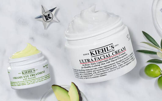 Alt-Text: Eine flache Anordnung von Kiehl's Hautpflegeprodukten auf einem marmorierten Hintergrund, umgeben von frischen Pflanzen und Zutaten.