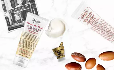 Eine Kiehl's Creme de Corps Tube, eine Gesichtsmaske und Argan-Nusse auf einem Tisch, umgeben von einer historischen Apotheke.