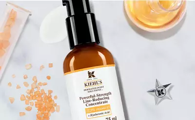 Braunes Serumflaschchen von Kiehl's, umgeben von Vitamin C-Kristallen und Hyaluronsaure, auf einem weißen Hintergrund.