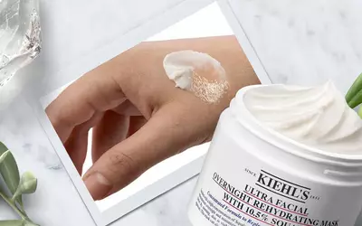 Eine Hand mit Kiehl's Ultra Facial Overnight Maske, daneben die offene Produktverpackung vor einem neutralen Hintergrund.