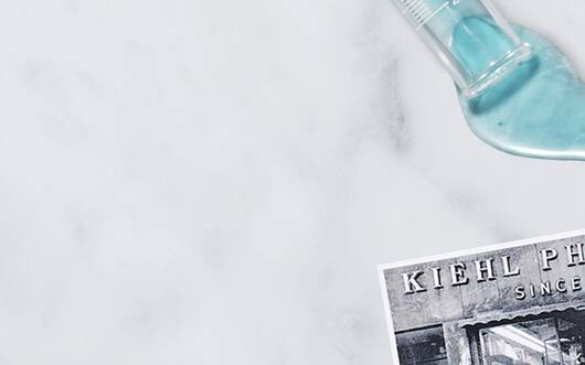 Alt-Text: Eine helle Arbeitsflache mit verschiedenen Kiehl's Hautpflegeprodukten, darunter ein Flaschchen mit Cannabis-Ol und einer blauen Lotion, umgeben von Kornern und frischen Blattern.