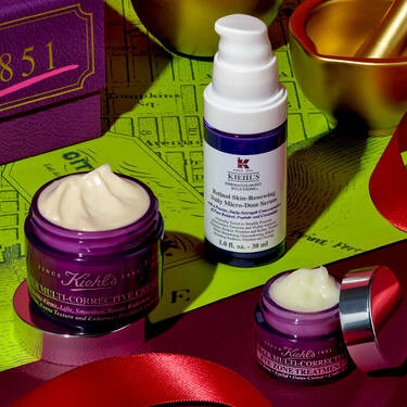 Drei Kiehl’s Super Multi-Corrective Produkte – ein Cremetiegel, eine Augencreme und eine Serumflasche – sind auf einem roten Band neben einer violetten Box und goldenen festlichen Dekorationen auf einem limettengrunen, landkartenartig gemusterten Hintergrund angeordnet.