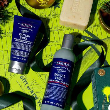 Kiehl’s Facial Fuel Cleanser und Feuchtigkeitspflege, arrangiert mit einer Seifenbar und gruner Weihnachtsdekoration auf einem leuchtend gelben Hintergrund.