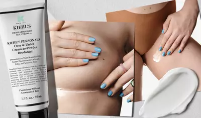 A tube of Kiehl's Over & Under Cream-to-Powder Deodorant is shown next to a close-up of a person applying the product under their arm. The hand has light blue nail polish and is gently smoothing the cream on the skin. A small amount of the cream is also displayed on a white surface.
